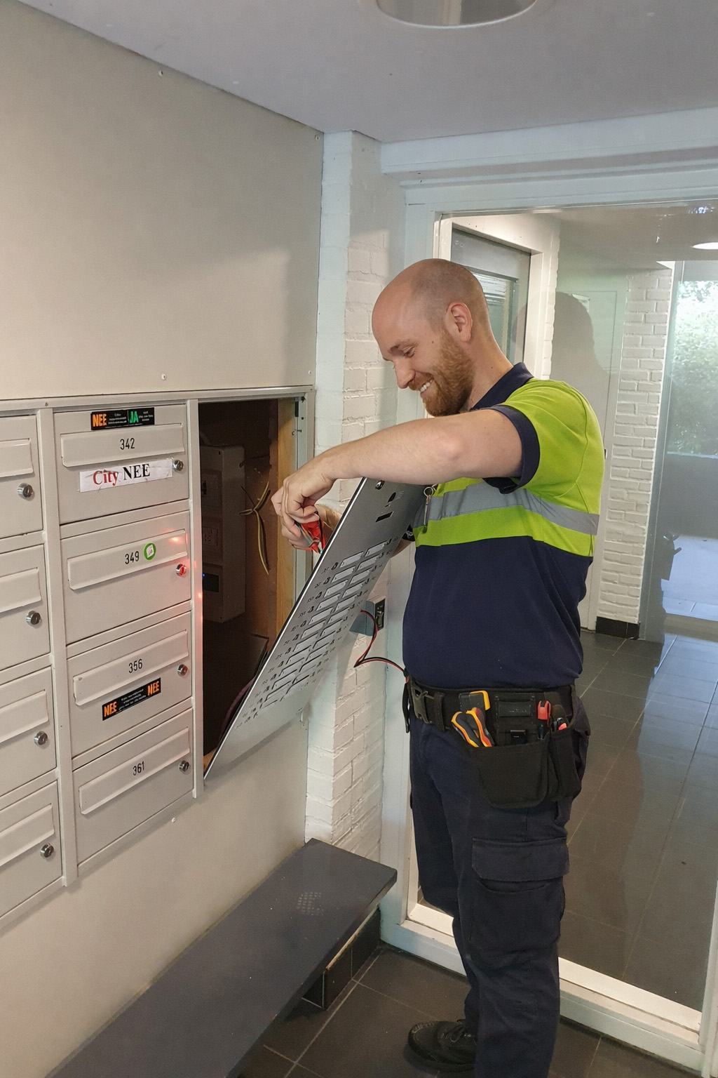 Een monteur is bezig met het openen en inspecteren van een intercom- of toegangscontrolesysteem dat is geïntegreerd in een wand met brievenbussen in een appartementencomplex. Hij houdt het frontpaneel open en werkt aan de interne bedrading met een gereedschap in de hand. De situatie speelt zich af in de entreehal van een woongebouw, waar de brievenbussen naast elkaar zijn gemonteerd en toegang wordt verleend via een centrale installatie.